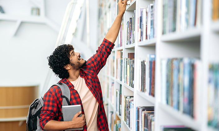 A male college student reaching for a book in the library to signify how are college scholarships and grants different?