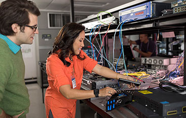 A systems security officer checks on hardware in a server room with a colleague